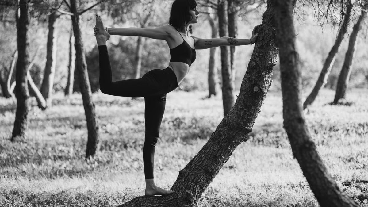Black and white side view of calm woman in sportswear doing Natarajasana near tree in nature